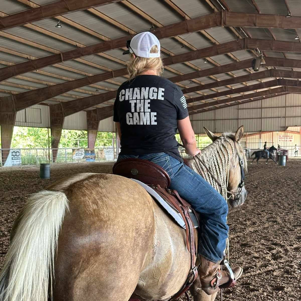 Person riding a horse in an indoor arena wearing a shirt with 'Change the Game' text.