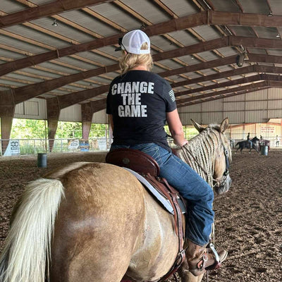 Person riding a horse in an indoor arena wearing a shirt with 'Change the Game' text.