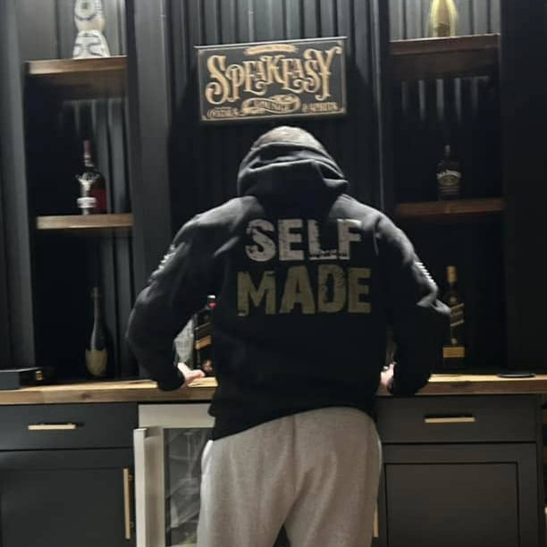Person wearing a hoodie with 'SELF MADE' text in a bar setting with Speakeasy sign.
