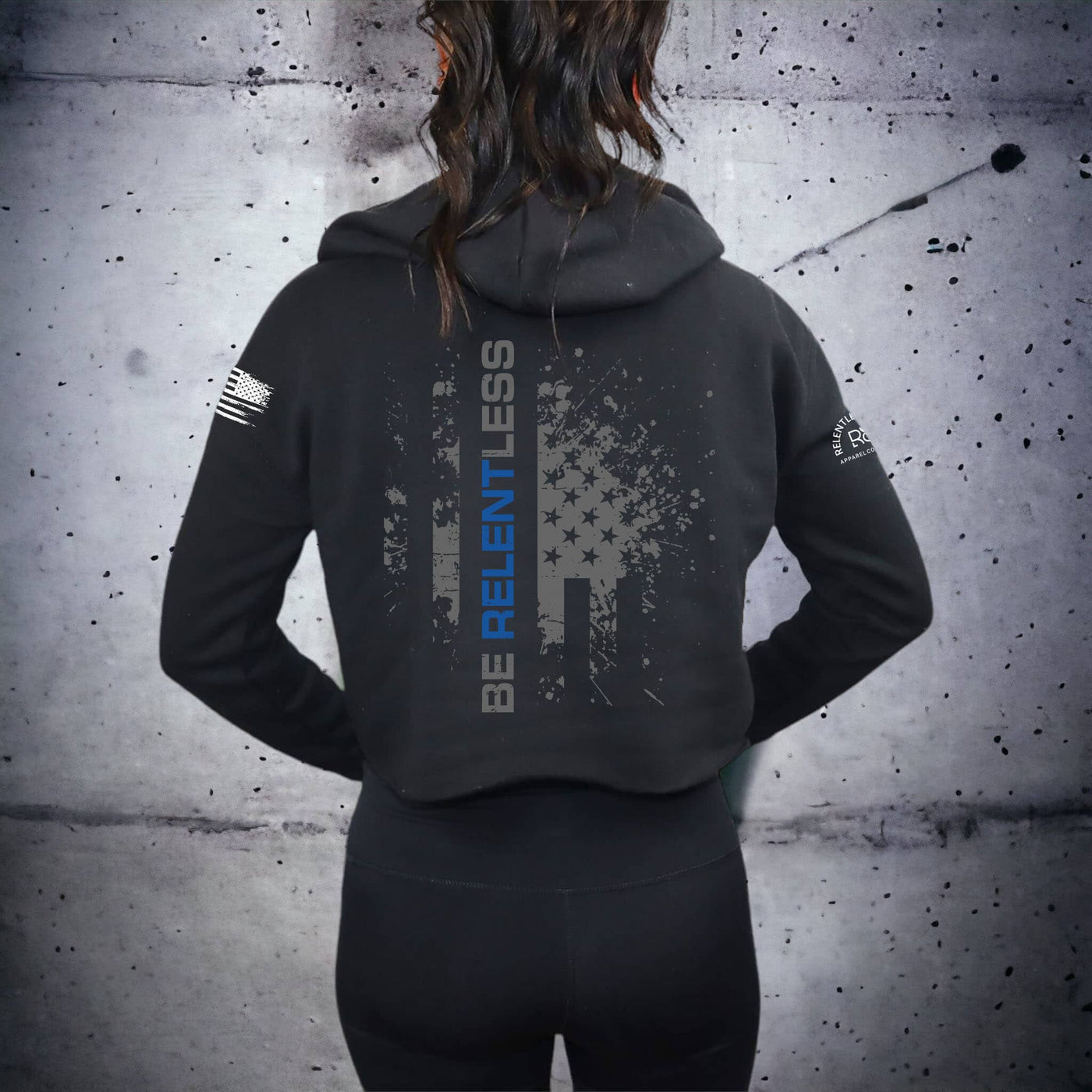 Person wearing a black hoodie with text and graphics on the back against a textured gray wall.