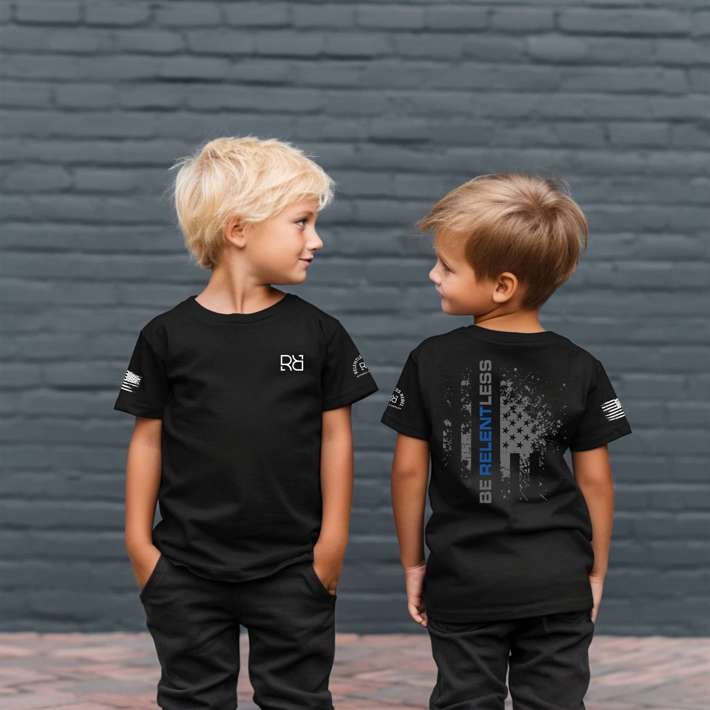Two children wearing black t-shirts with visible branding against a brick wall.