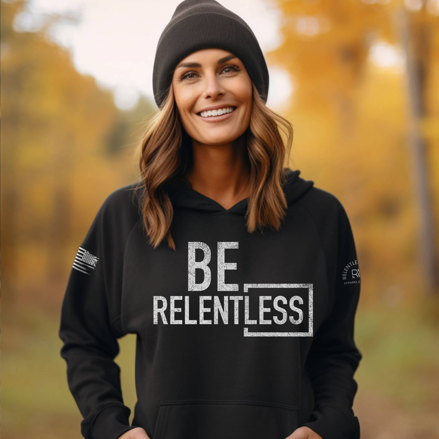 Woman wearing a black hoodie with 'BE RELENTLESS' text in an autumn forest setting