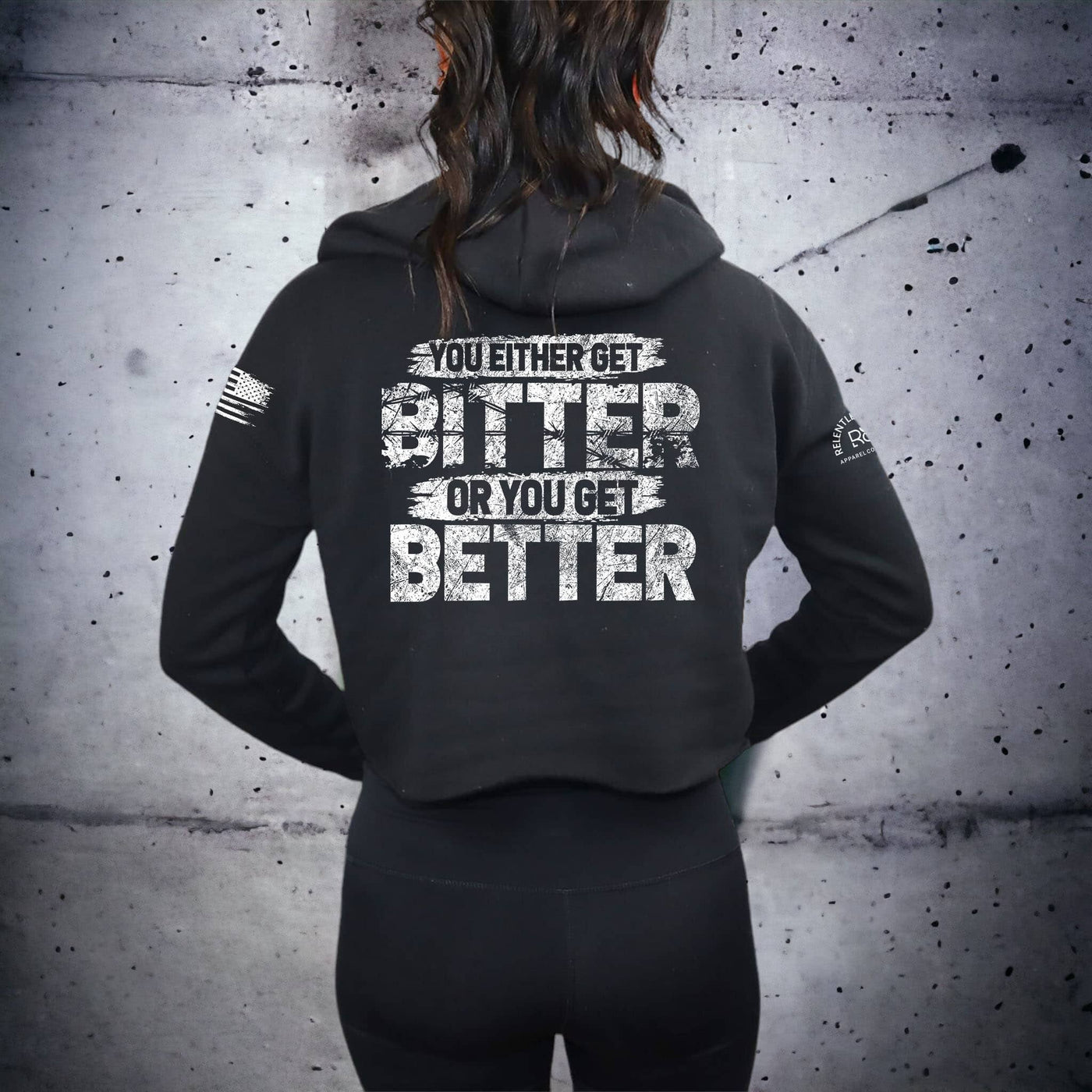 Person wearing a black hoodie with motivational text on the back against a gray concrete wall.