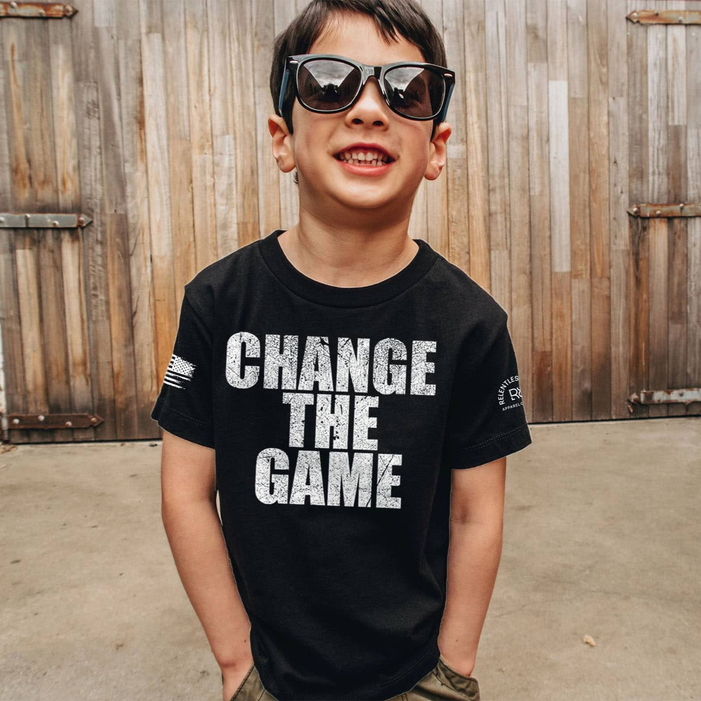 Child wearing a black t-shirt with 'CHANGE THE GAME' text in front of wooden shutters.
