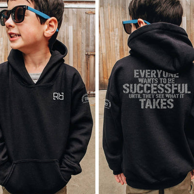 Child wearing a black hoodie with a logo on the front and motivational text on the back, standing in front of a wooden fence.