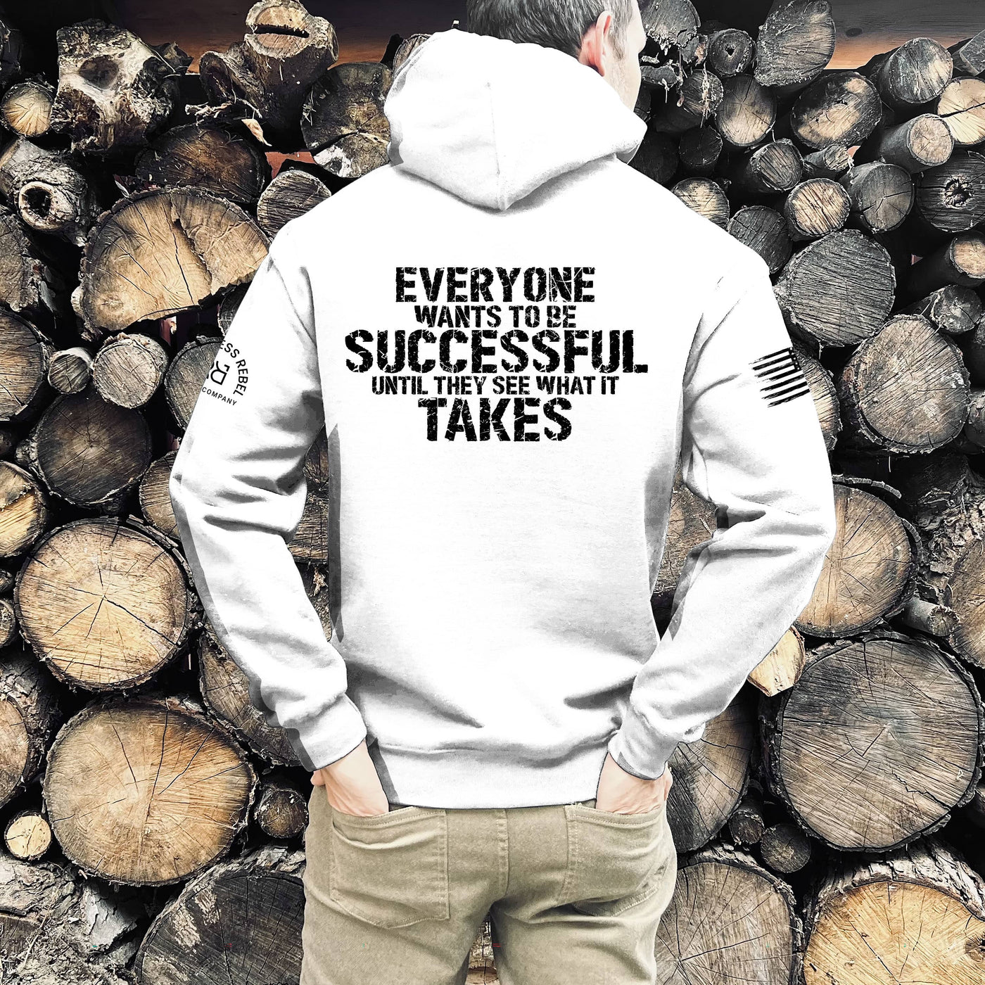 Person wearing a white hoodie with a motivational quote on the back, standing against a background of stacked wooden logs.