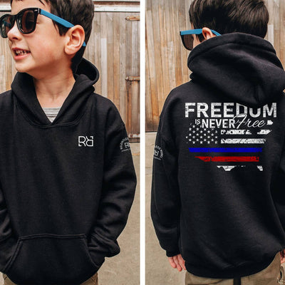 Child wearing a black hoodie with 'Freedom Never Free' design and sunglasses, standing in front of a wooden fence.