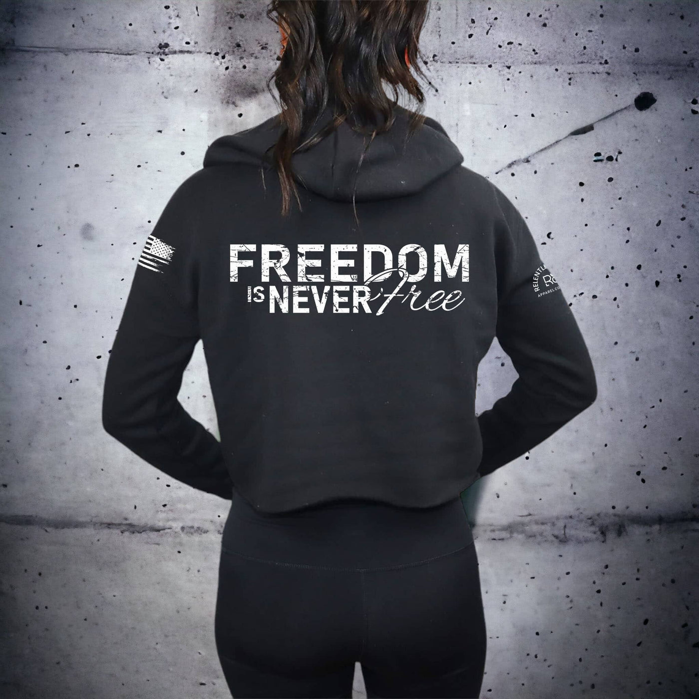 Person wearing a black hoodie with 'Freedom is Never Free' text on the back against a textured wall.