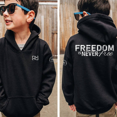 Child wearing a black hoodie with 'Freedom is Never Free' text on the back, standing in front of a wooden fence.