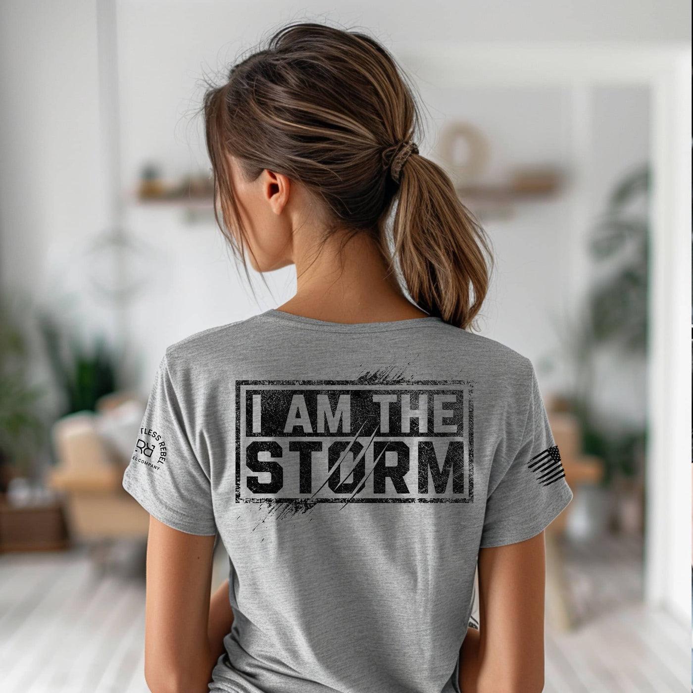 Person wearing a gray t-shirt with 'I AM THE STORM' text on the back in a blurred indoor setting.