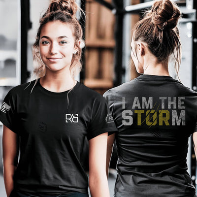 Woman wearing a black t-shirt with a logo on the front and 'I AM THE STORM' text on the back, standing in a gym setting.