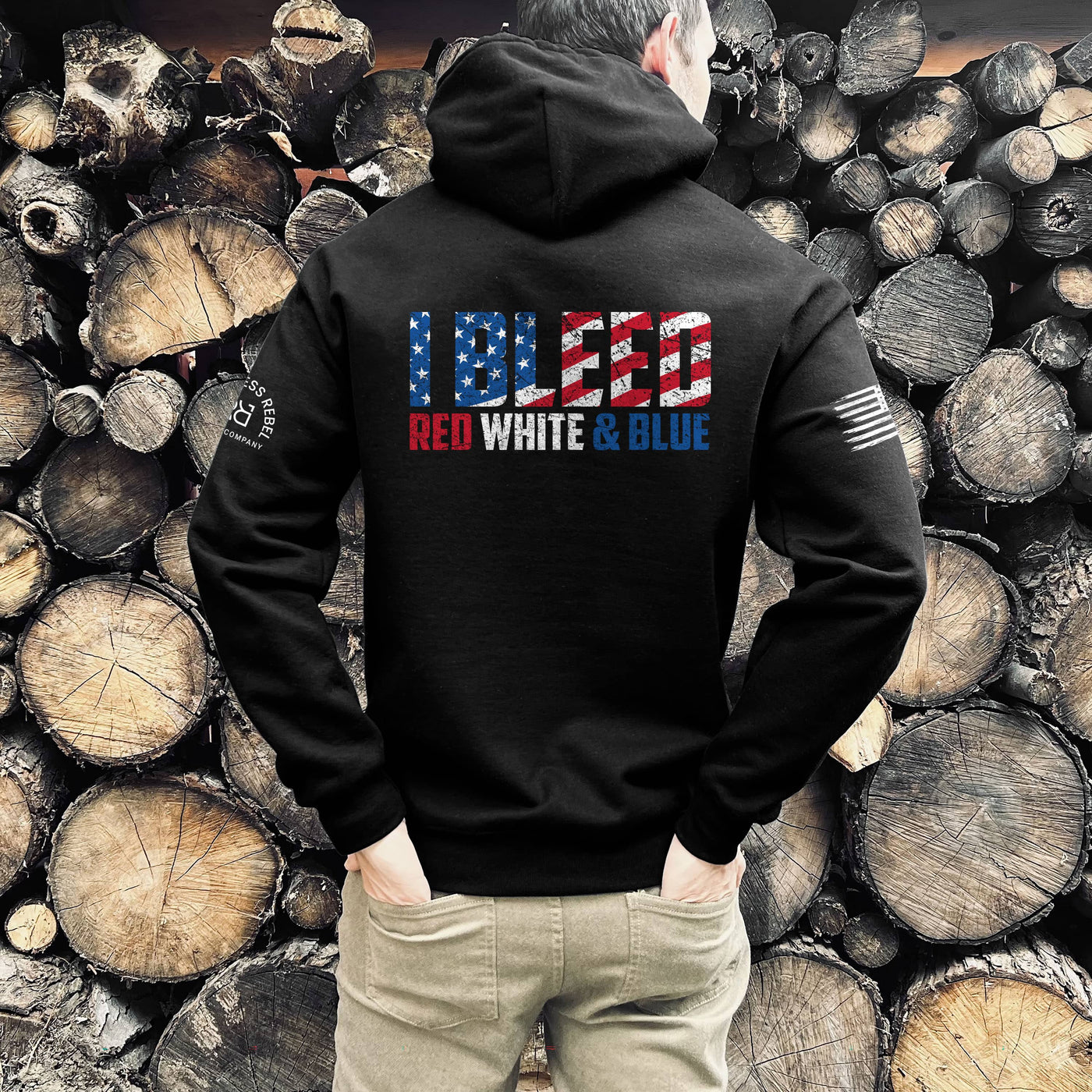 Person wearing a black hoodie with 'I BLEED RED WHITE & BLUE' text in front of stacked wooden logs.