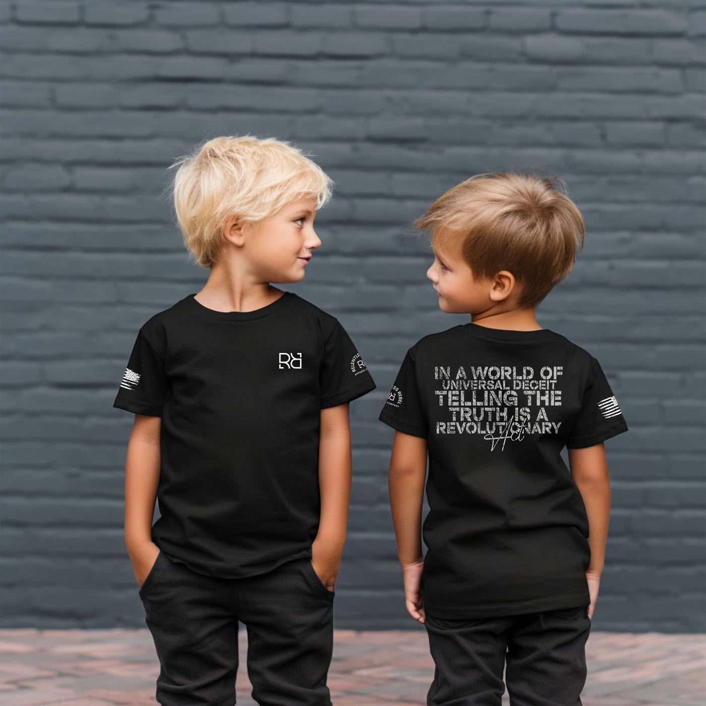 Two children wearing black t-shirts with text against a brick wall.