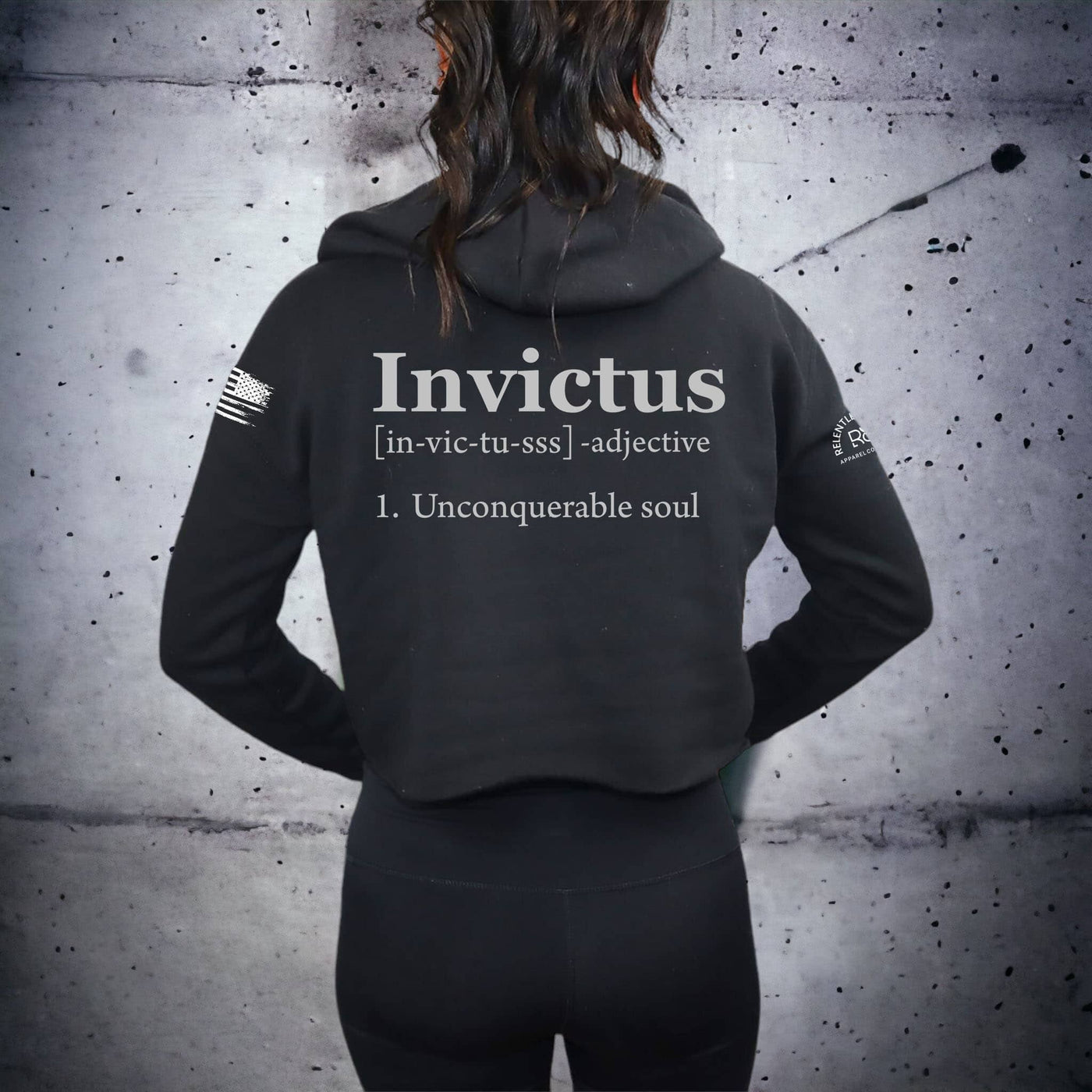 Person wearing a black hoodie with 'Invictus' text on the back against a textured wall.