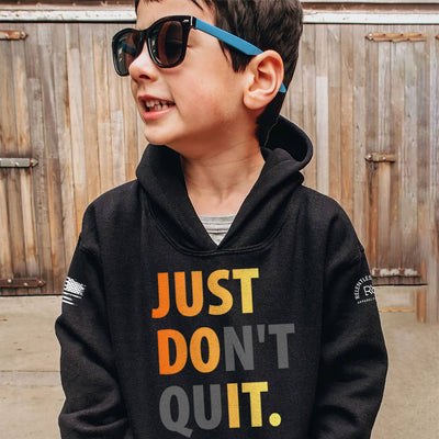 Child wearing a black hoodie with 'JUST DON'T QUIT.' text, standing in front of a wooden fence.