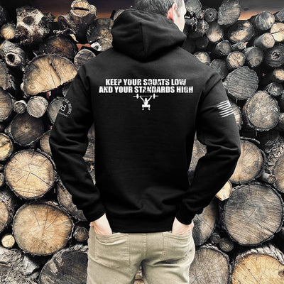 Person wearing a black hoodie with text and graphics, standing against a background of stacked wooden logs.