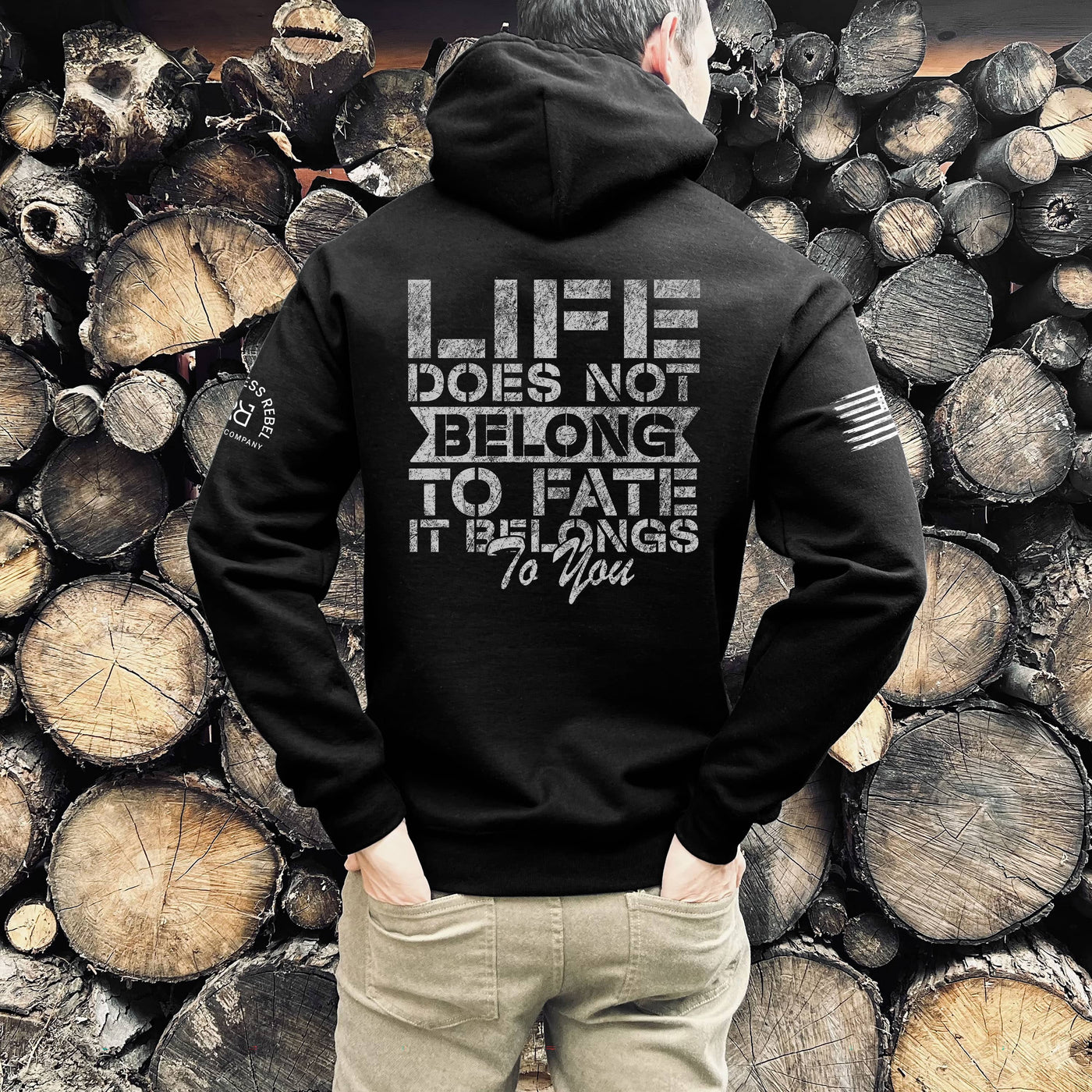 Person wearing a black hoodie with a motivational quote on the back, standing against a background of stacked wooden logs.