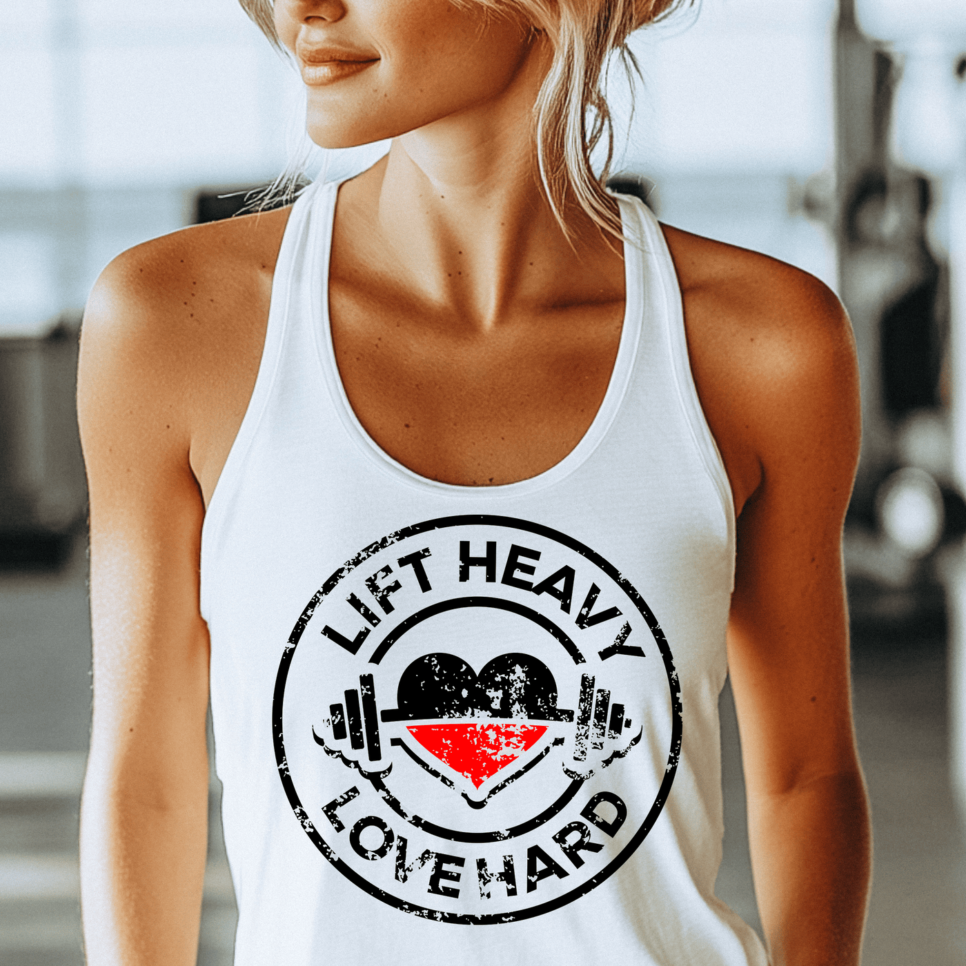 Woman wearing a white tank top with 'Lift Heavy Love Hard' graphic in a gym setting