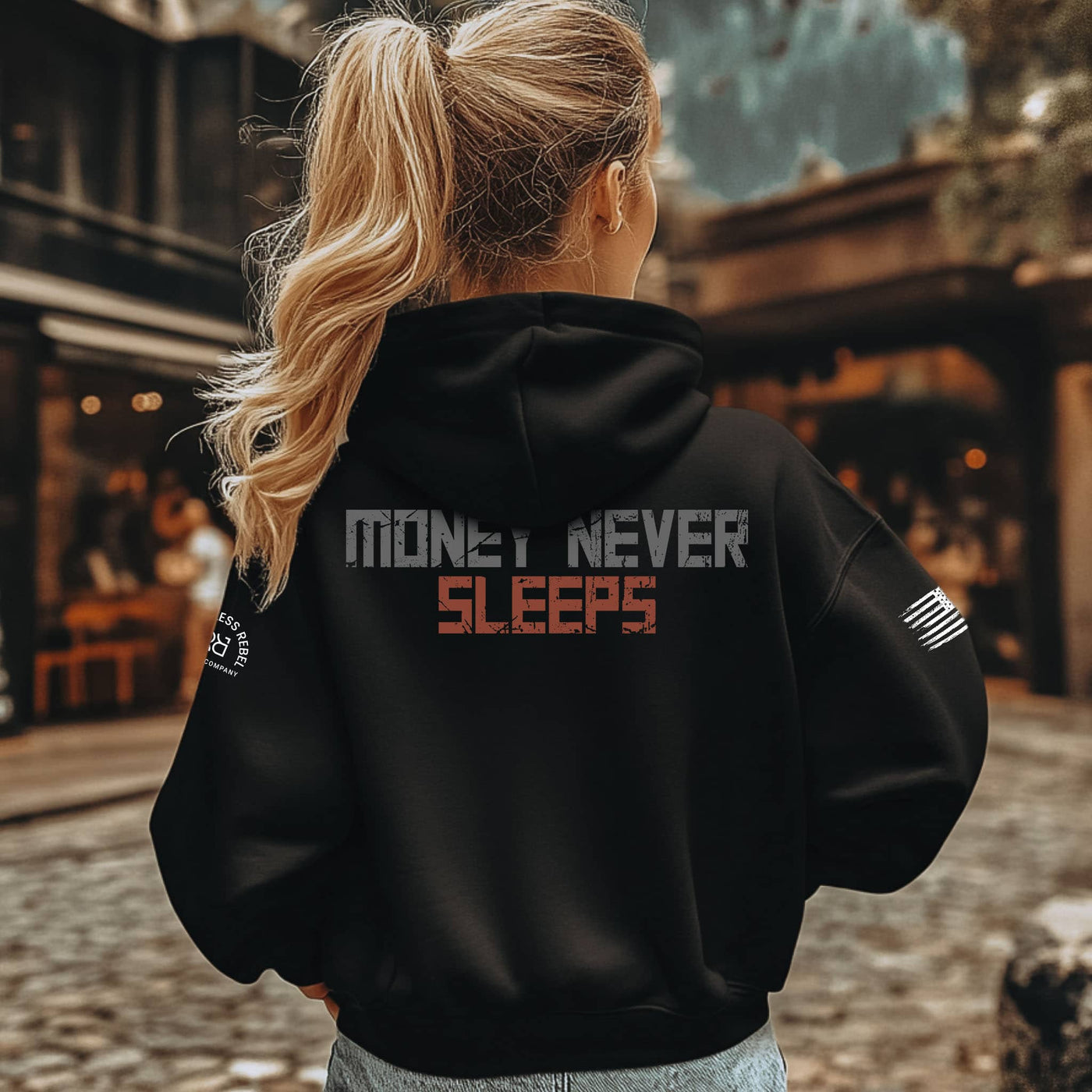 Person wearing a black hoodie with 'Money Never Sleeps' text on the back, standing outdoors.