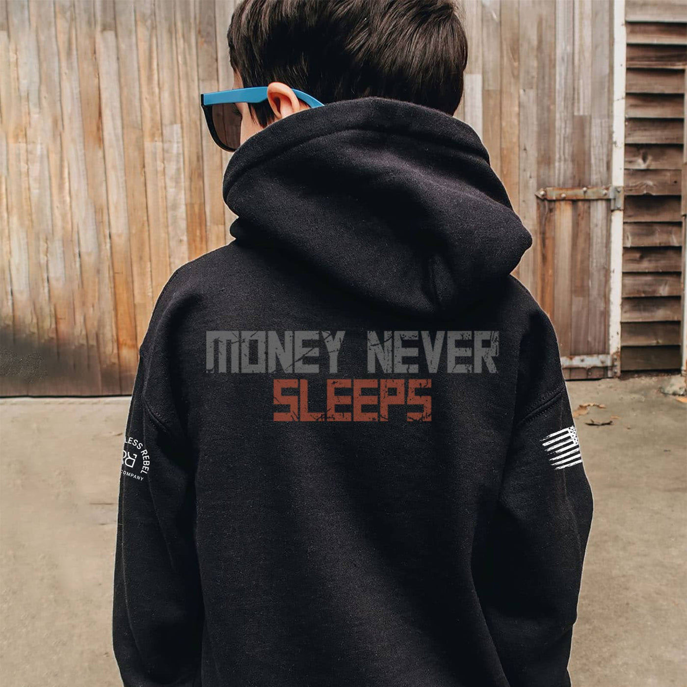 Person wearing a black hoodie with 'Money Never Sleeps' text on the back, standing in front of a wooden wall.