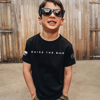 Child wearing a black t-shirt with 'Raise The Bar' text in front of wooden shutters.