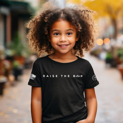 Child wearing a black t-shirt with 'Raise the Bar' text in an outdoor setting