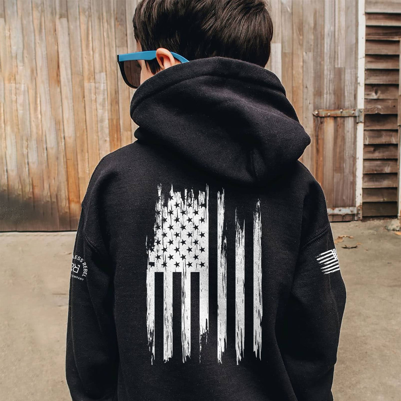 Person wearing a black hoodie with a white flag design on the back, standing in front of a wooden wall.