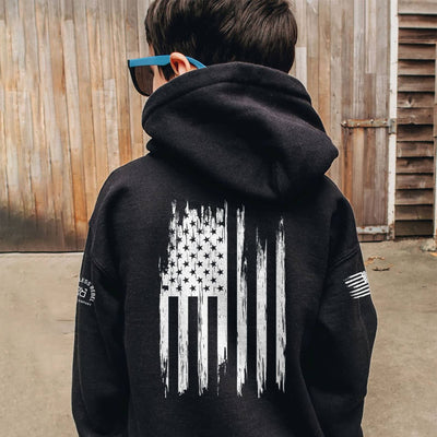Person wearing a black hoodie with a white flag design on the back, standing in front of a wooden wall.