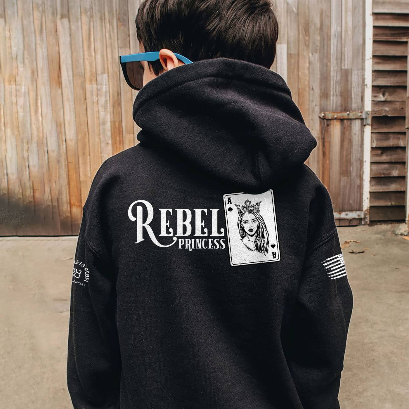 Person wearing a black hoodie with 'Rebel Princess' design on the back, standing in front of wooden panels.