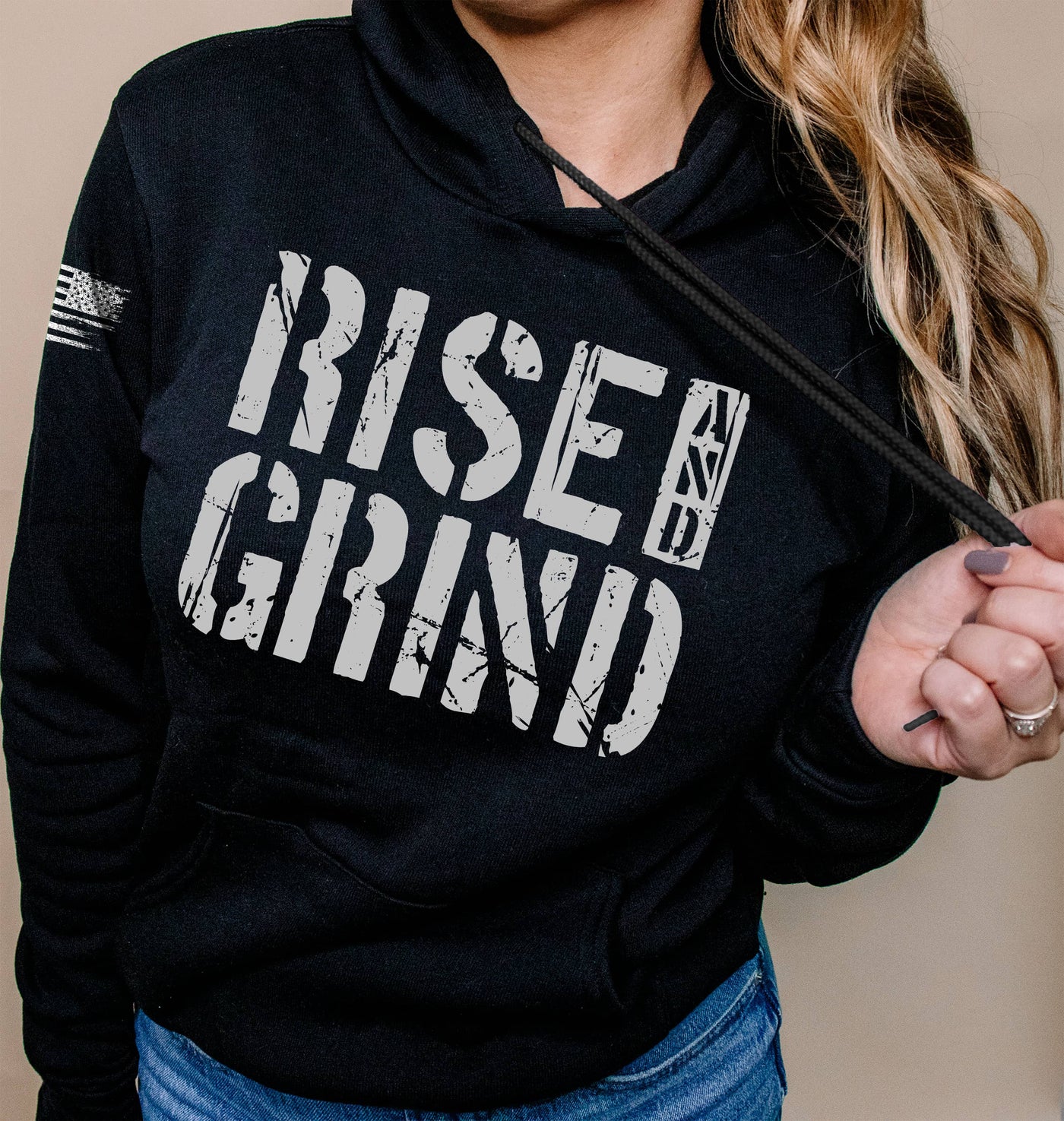 Person wearing a black hoodie with 'Rise & Grind' text on a beige background