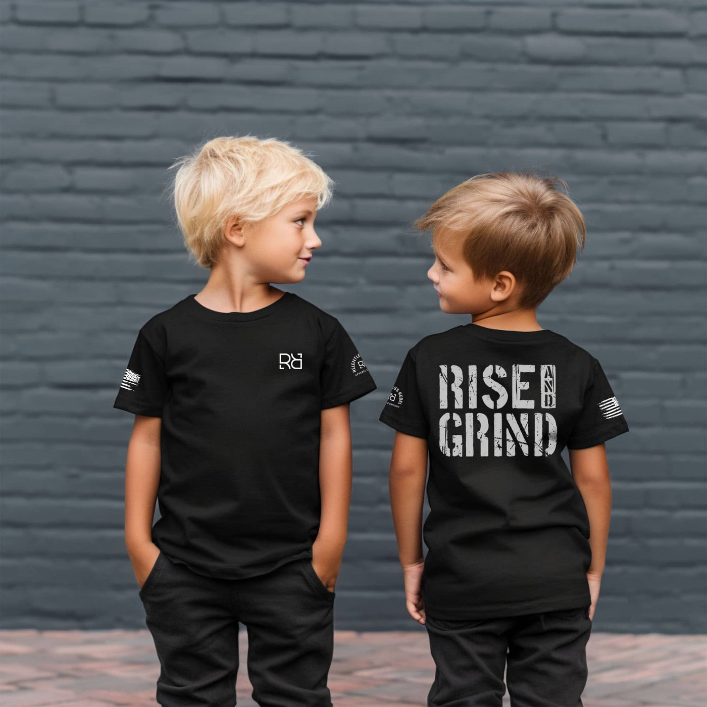 Two children wearing black t-shirts with 'Rise & Grind' text against a brick wall.
