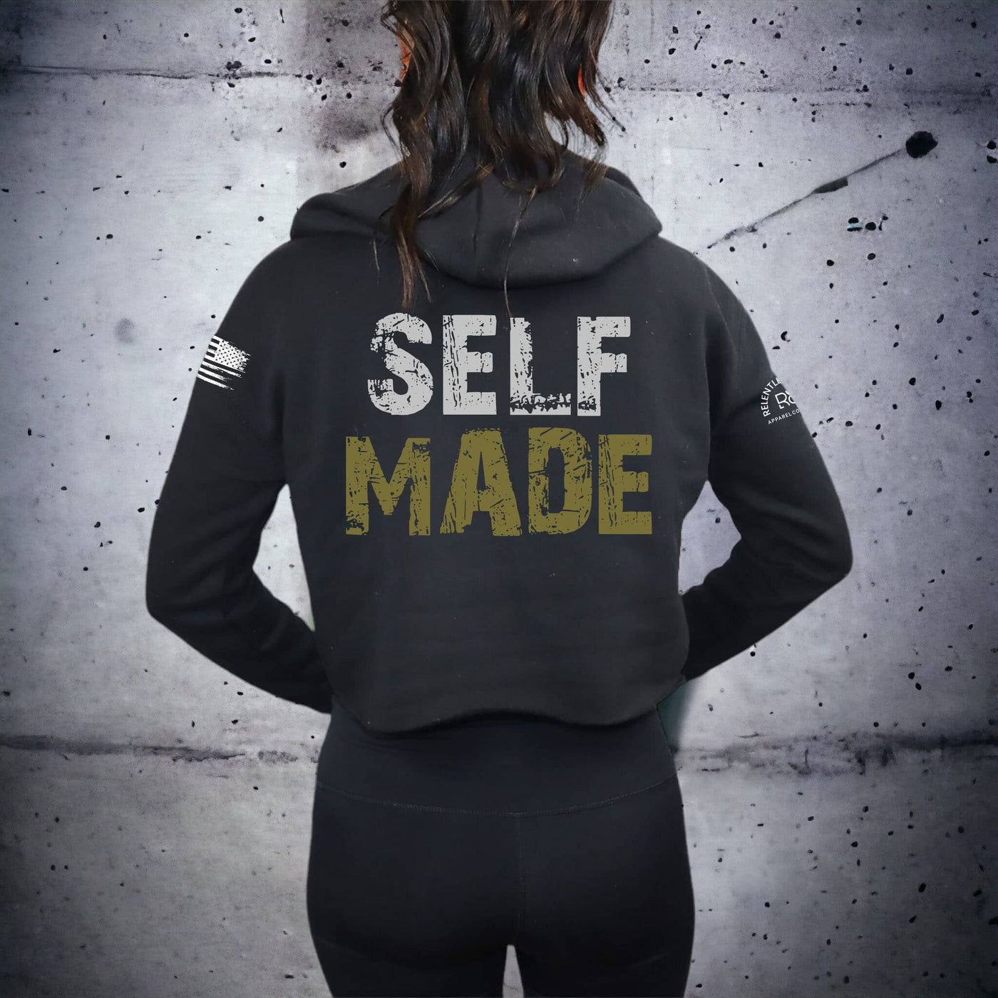 Person wearing a black hoodie with 'SELF MADE' text on the back against a textured gray wall.