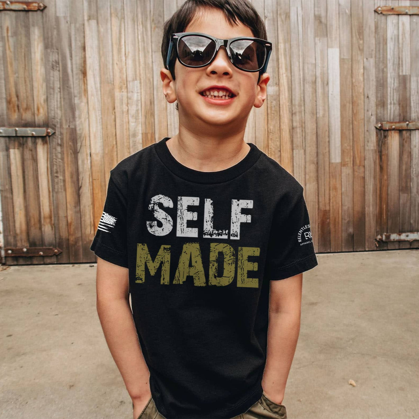 Child wearing a black t-shirt with 'SELF MADE' text, sunglasses, standing in front of wooden shutters.