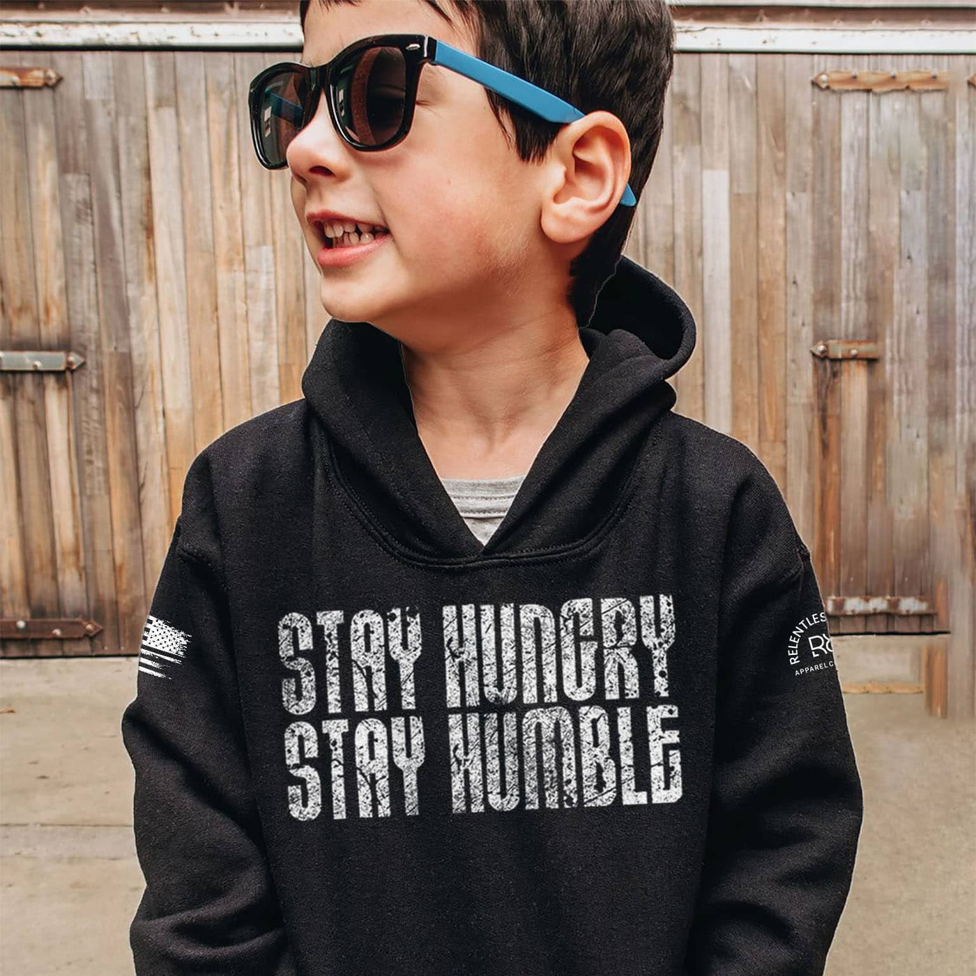 Child wearing a black hoodie with 'Stay Hungry Stay Humble' text, standing in front of a wooden fence.