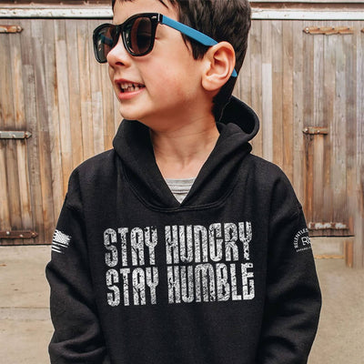 Child wearing a black hoodie with 'Stay Hungry Stay Humble' text, standing in front of a wooden fence.