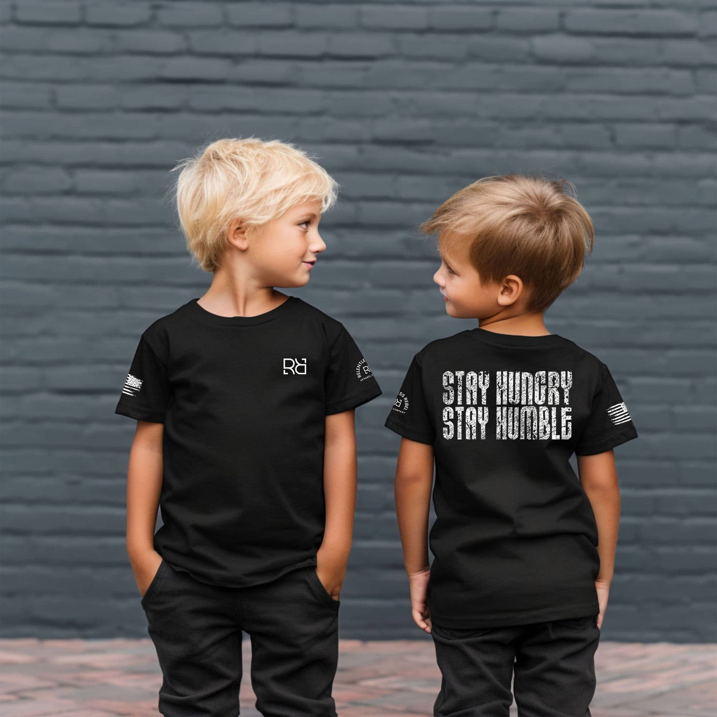 Two children wearing black t-shirts with text against a brick wall.