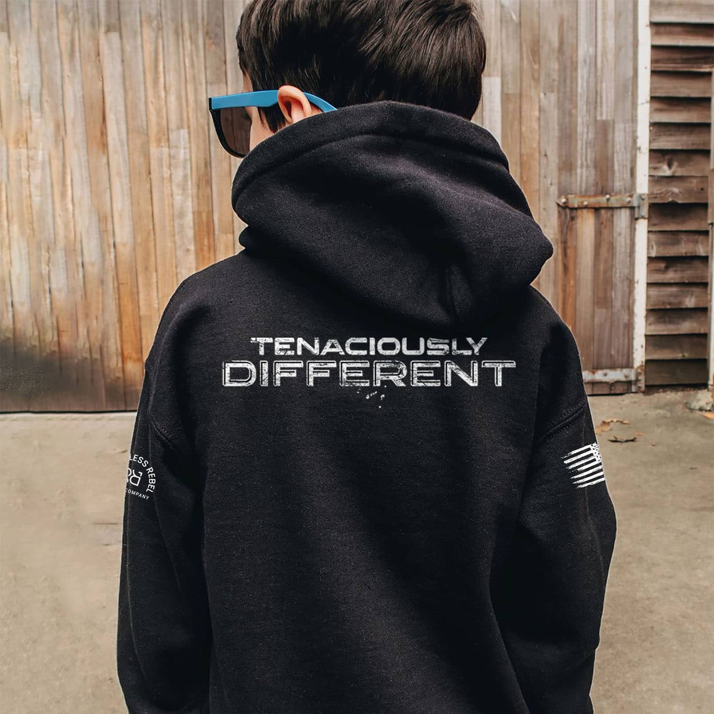 Person wearing a black hoodie with 'Tenaciously Different' text on the back, standing in front of a wooden wall.
