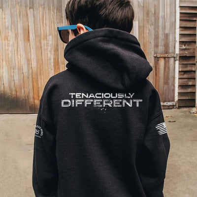 Person wearing a black hoodie with 'Tenaciously Different' text on the back, standing in front of a wooden wall.