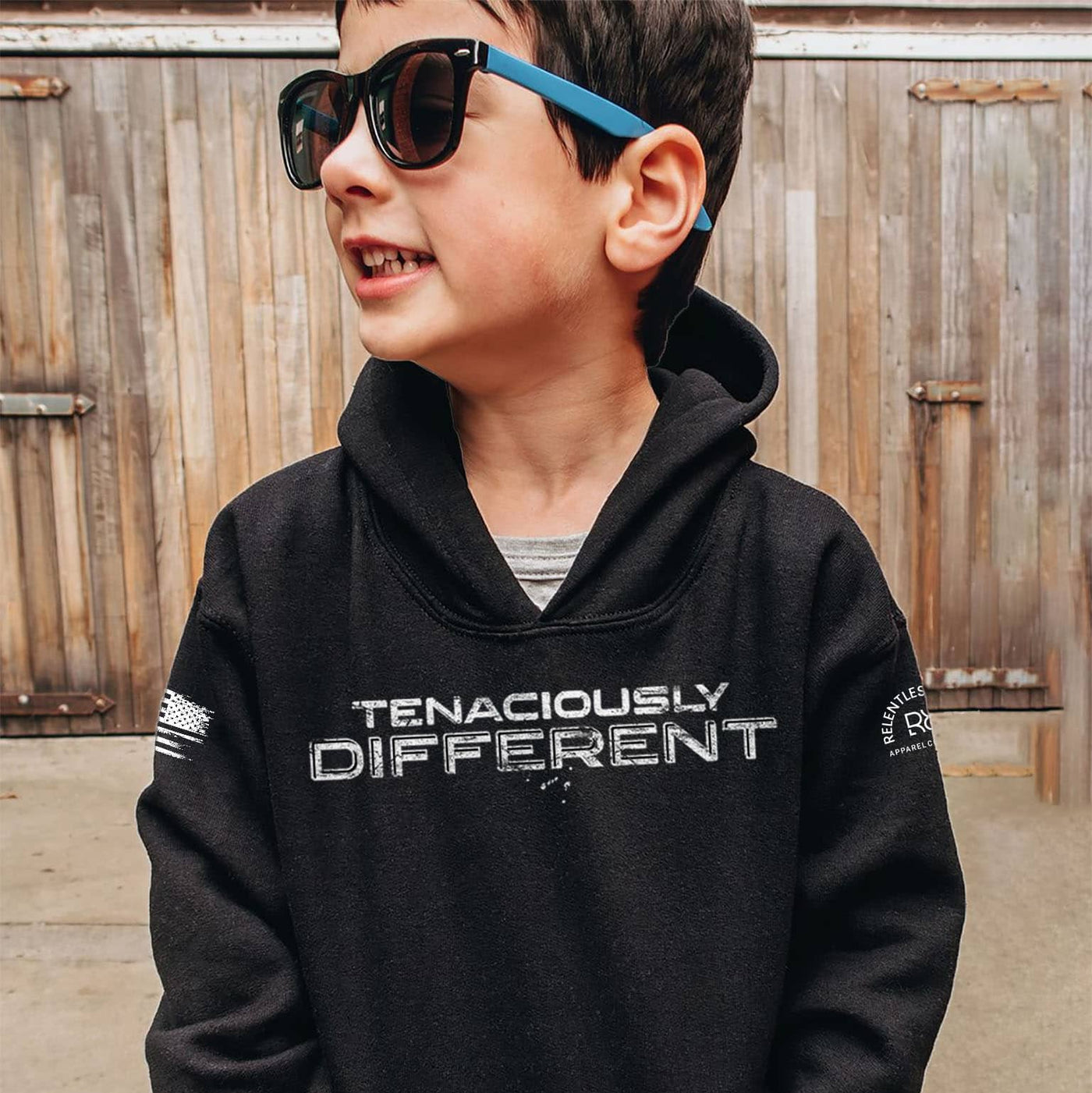 Child wearing a black hoodie with 'TENACIOUSLY DIFFERENT' text, standing in front of a wooden fence.