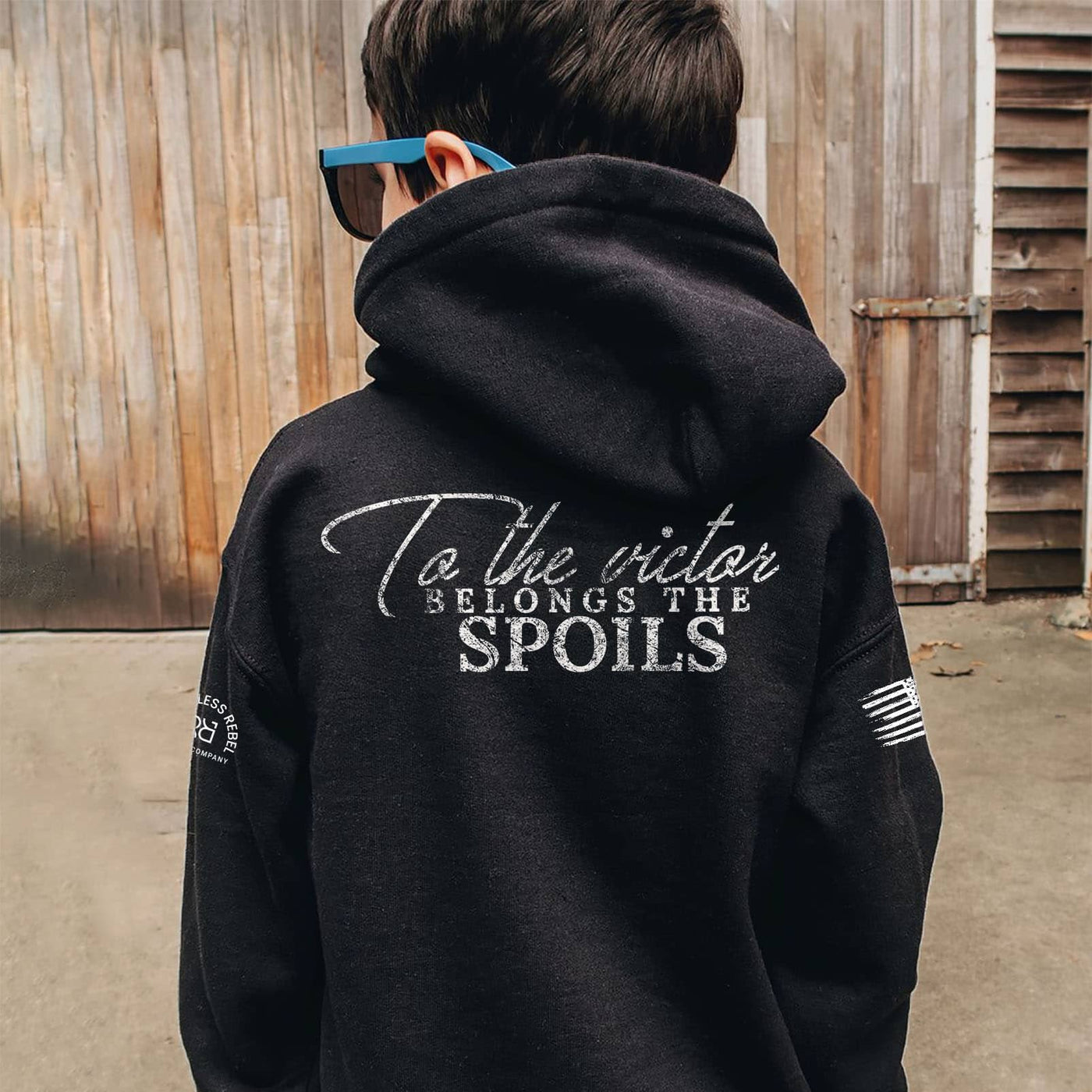 Person wearing a black hoodie with text on the back, standing in front of a wooden wall.