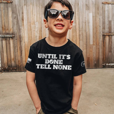 Child wearing a black t-shirt with text, standing in front of wooden shutters.