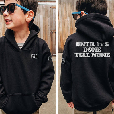 Child wearing a black hoodie with a logo on the front and text on the back, standing in front of a wooden fence.