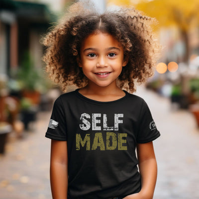 Child wearing a black t-shirt with 'SELF MADE' text, standing outdoors.
