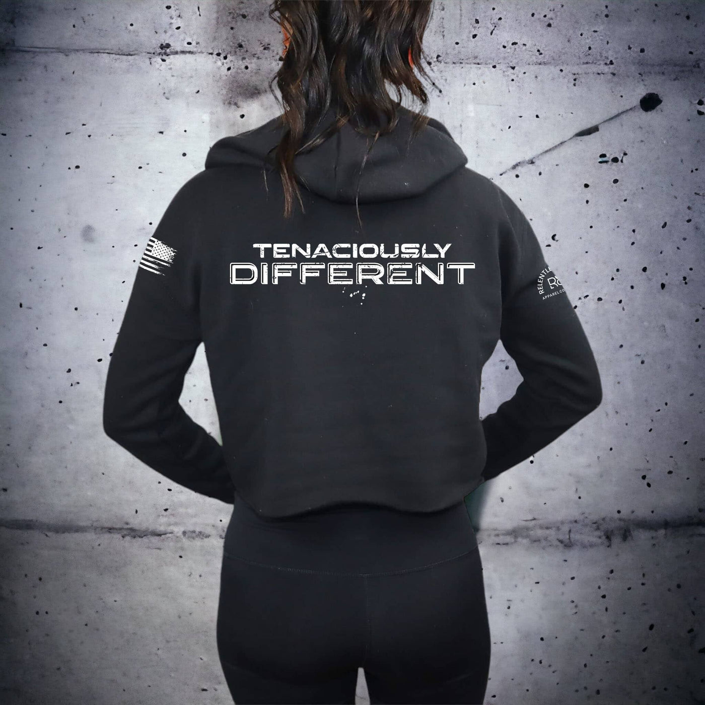 Person wearing a black hoodie with 'Tenaciously Different' text on the back against a textured gray wall.