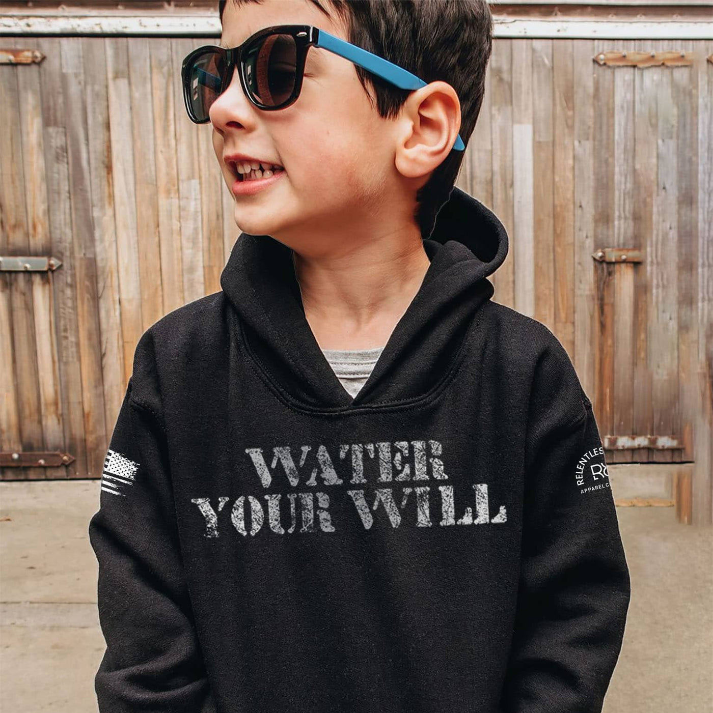 Child wearing a black hoodie with 'Water Your Will' text, standing in front of a wooden fence.