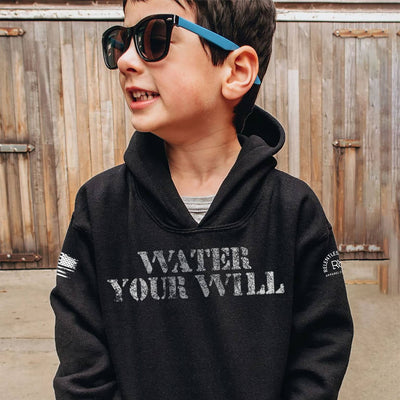 Child wearing a black hoodie with 'Water Your Will' text, standing in front of a wooden fence.