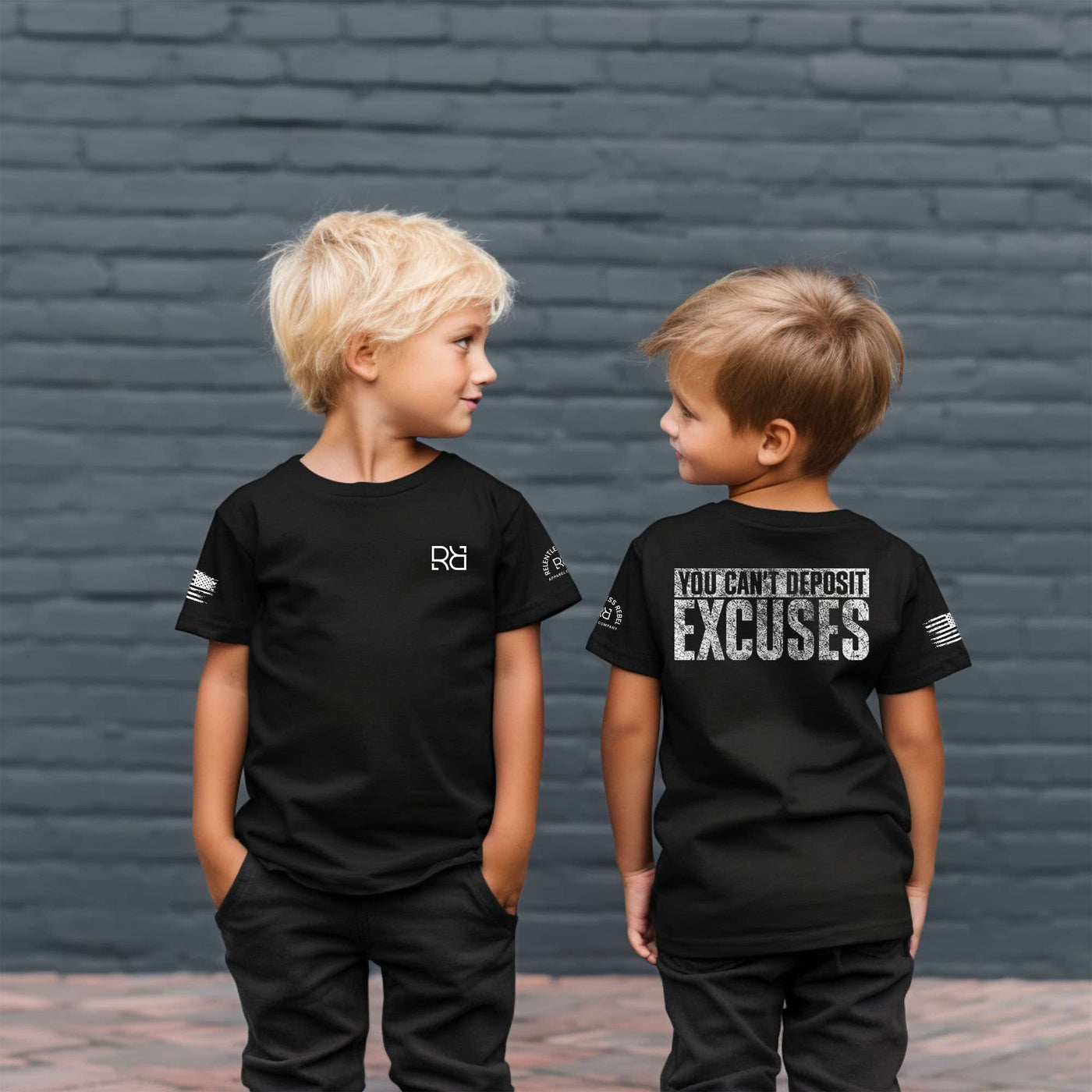 Two children wearing black t-shirts with text and logos against a brick wall.
