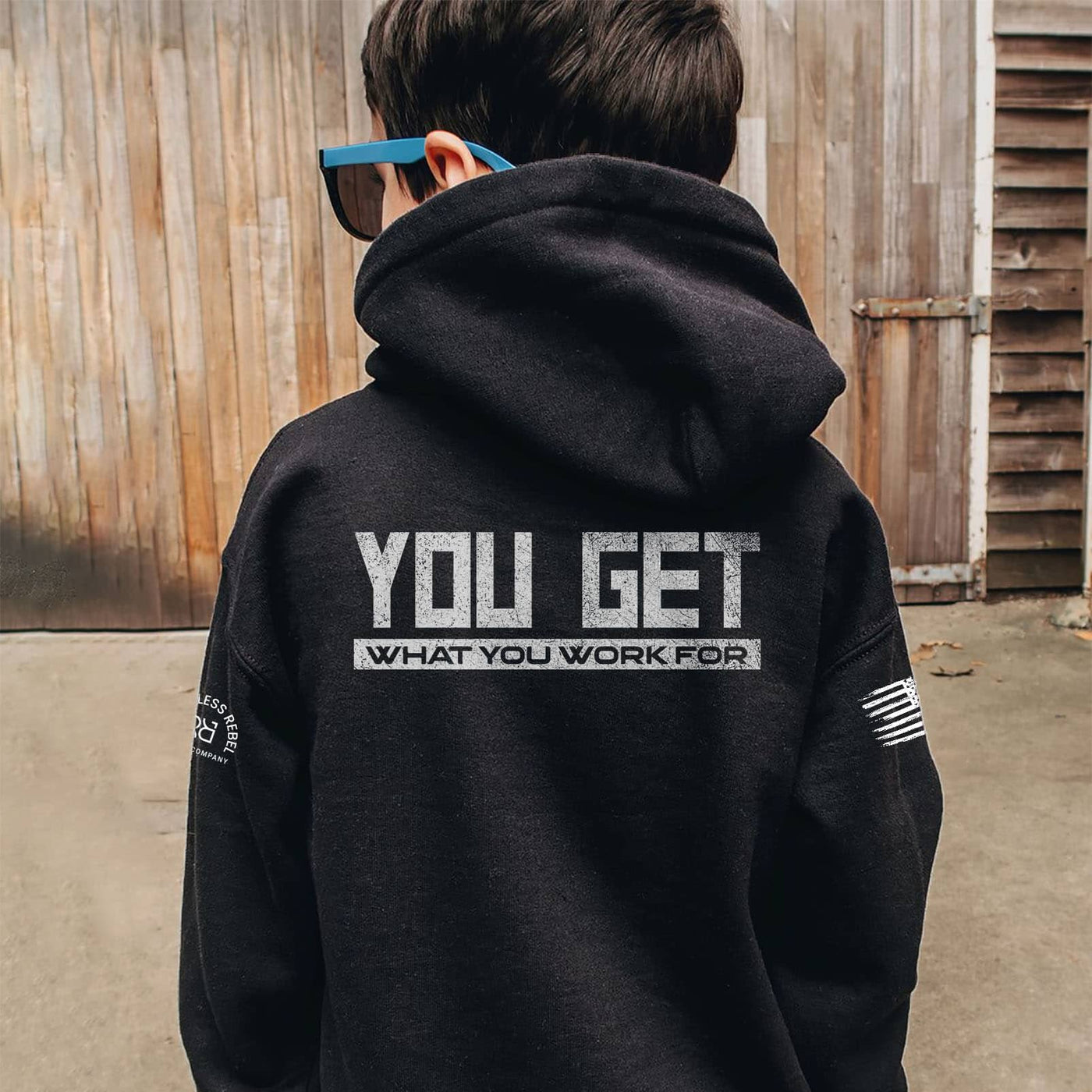 Person wearing a black hoodie with text on the back, standing in front of a wooden wall.