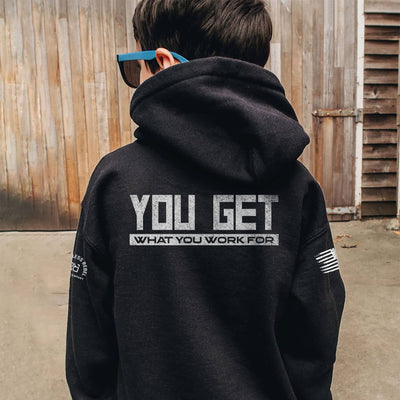 Person wearing a black hoodie with text on the back, standing in front of a wooden wall.