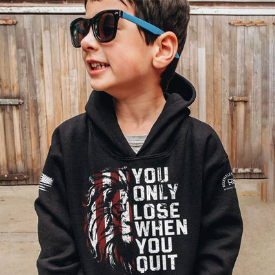 Child wearing a black hoodie with motivational quote and sunglasses, standing against a wooden fence.